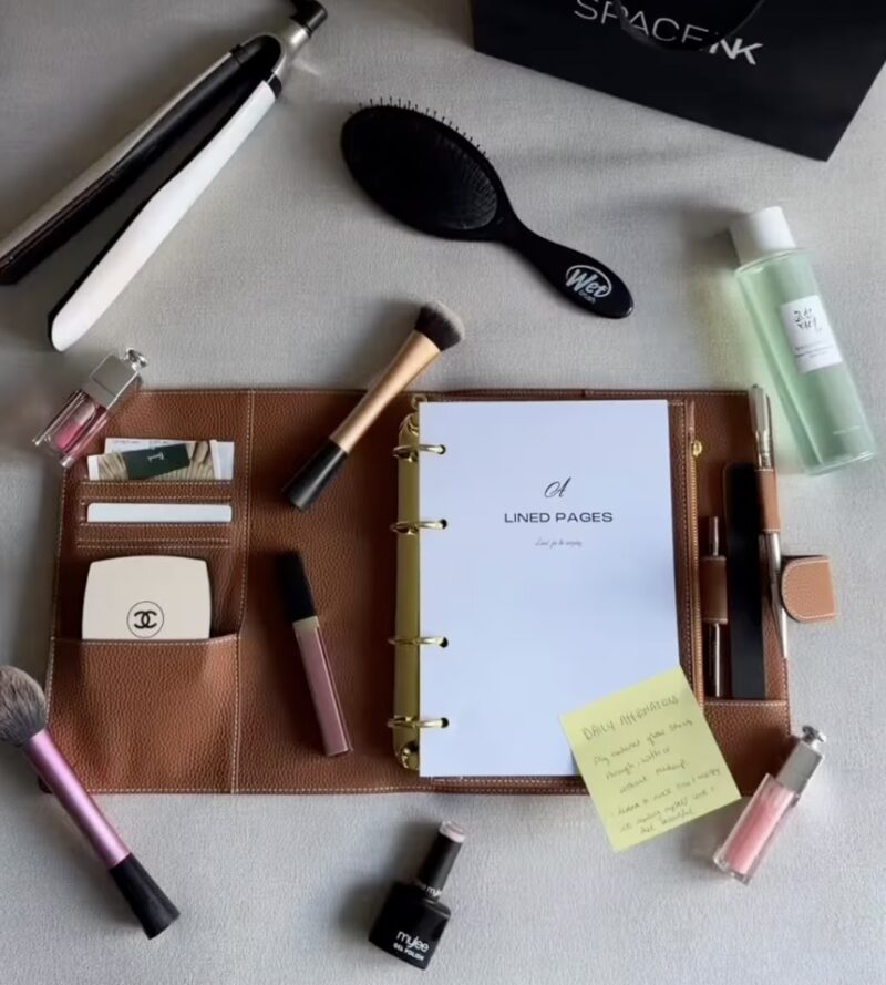 Geöffneter brauner Agende-Ringplaner mit Papier-Einlagen, Karten und Kosmetik, umgeben von Beauty-Produkten.